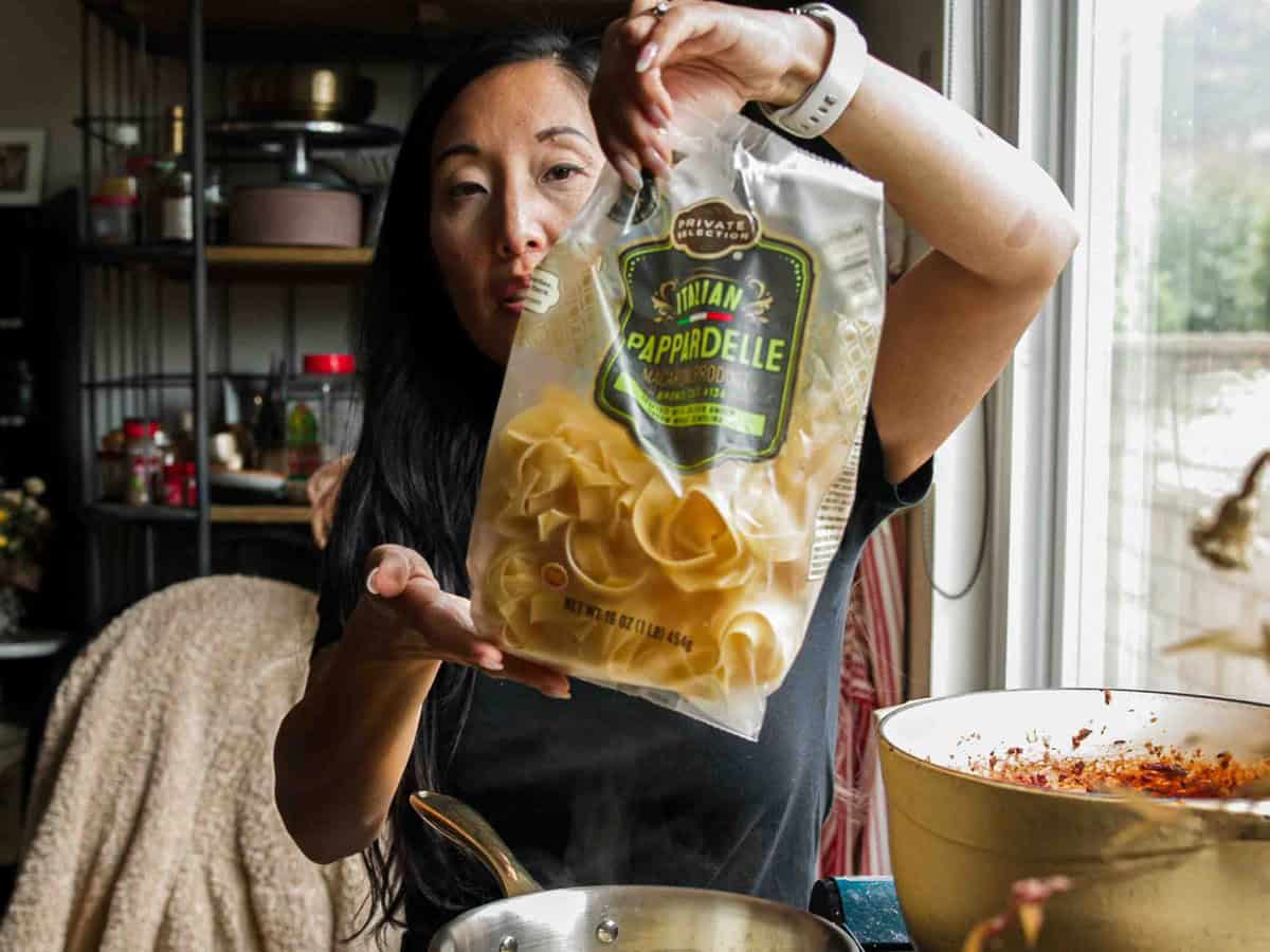A woman in a kitchen holds up a bag of Private Selection Pappardelle pasta over a stovetop with a pot of sauce and a pan, preparing to cook.